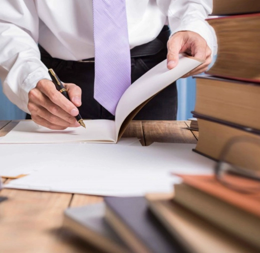 Primer plano de un profesional en camisa y corbata escribiendo con una pluma mientras revisa documentos o un libro, con una pila de libros sobre el escritorio.
