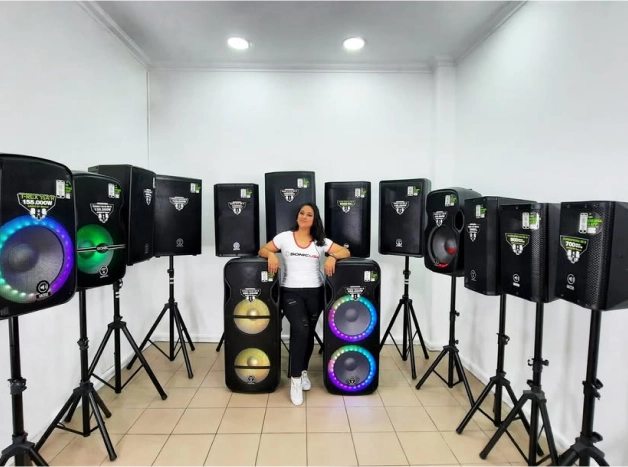 Una mujer sonriente posa en una tienda rodeada de múltiples altavoces de audio grandes de diferentes tamaños, incluyendo dos modelos coloridos en primer plano.